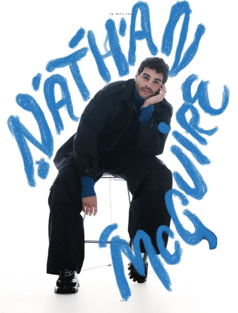 Portrait of a young man wearing a dark jacket and pants, seated on a stool against a white backdrop with blue text overlay.