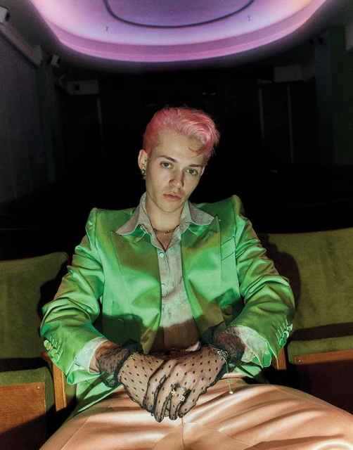 Stylish young man with pink hair in a lime green suit sitting in a vintage theatre with dark seating and a pink-lit ceiling.