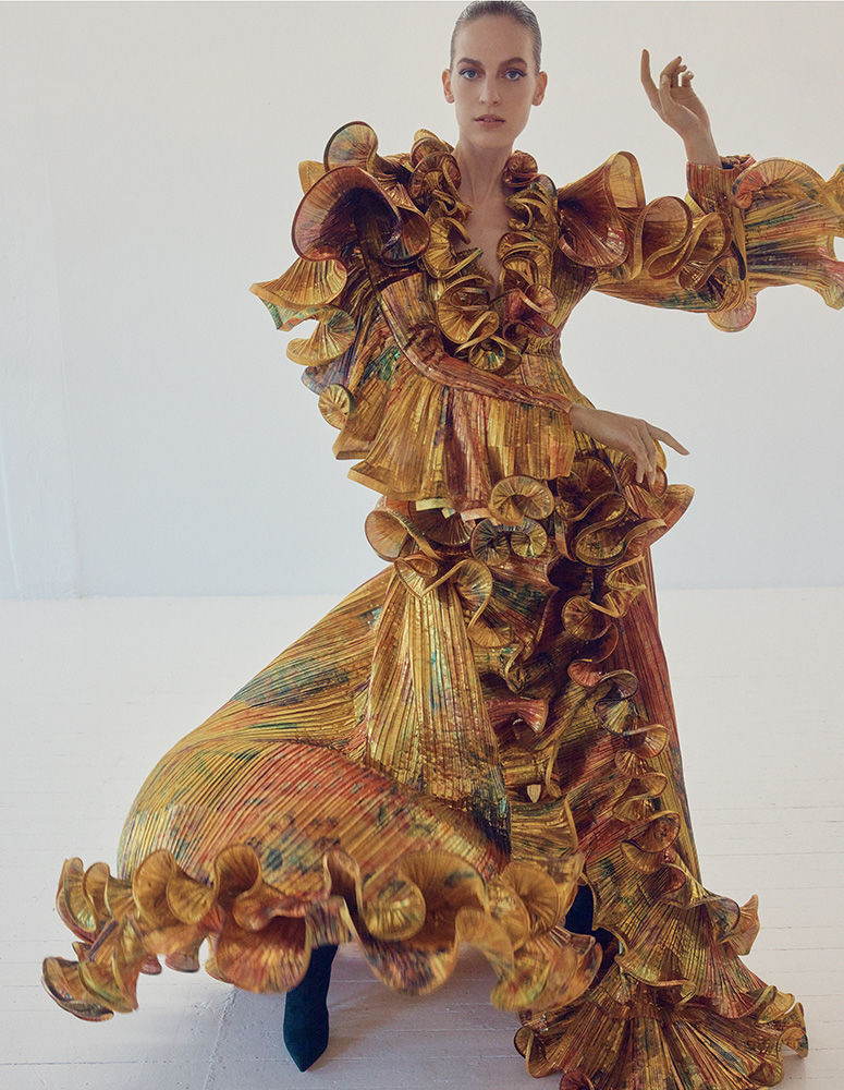 Adult female posing in a ruffled gold iridescent dress against a white background in a studio. The woman has her right arm raised.