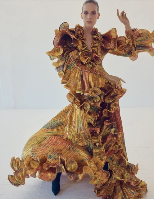 Adult female posing in a ruffled gold iridescent dress against a white background in a studio. The woman has her right arm raised.