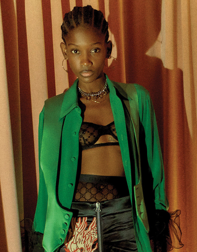 Portrait of a young adult woman with braided hair wearing a green satin jacket, black bra, and patterned pants in a studio setting.