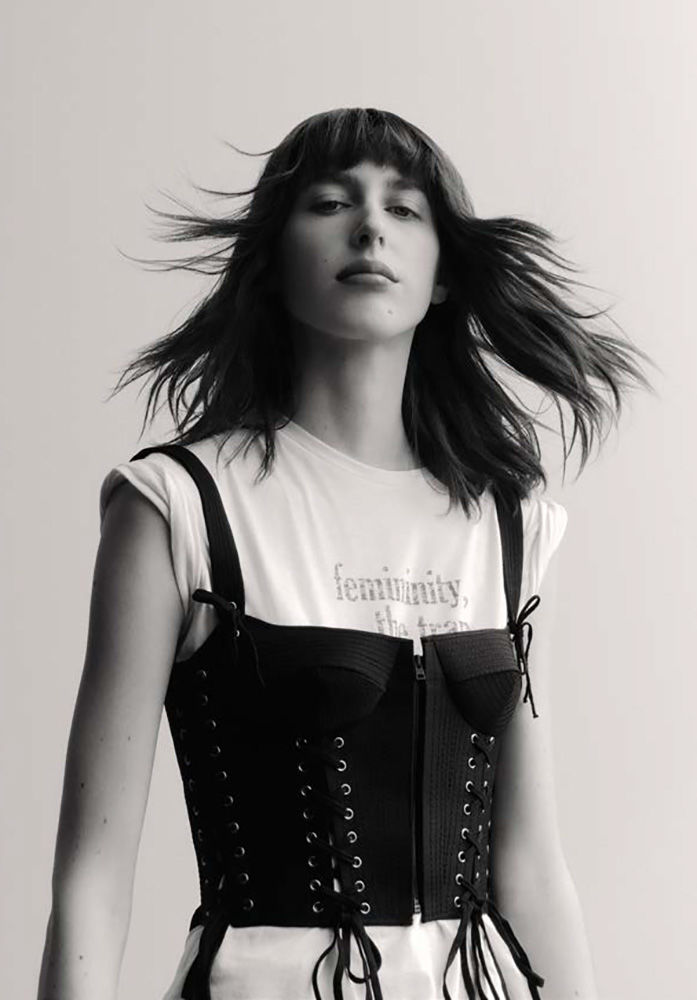 Monochrome portrait of a woman with dark hair and bangs wearing a laced black corset over a white t-shirt against a light background