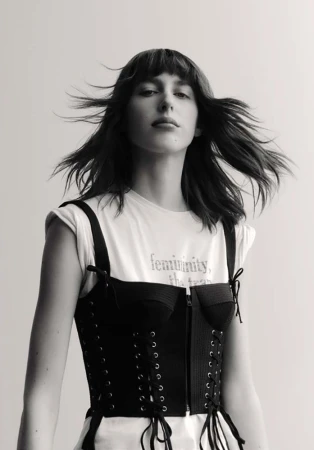 Monochrome portrait of a woman with dark hair and bangs wearing a laced black corset over a white t-shirt against a light background