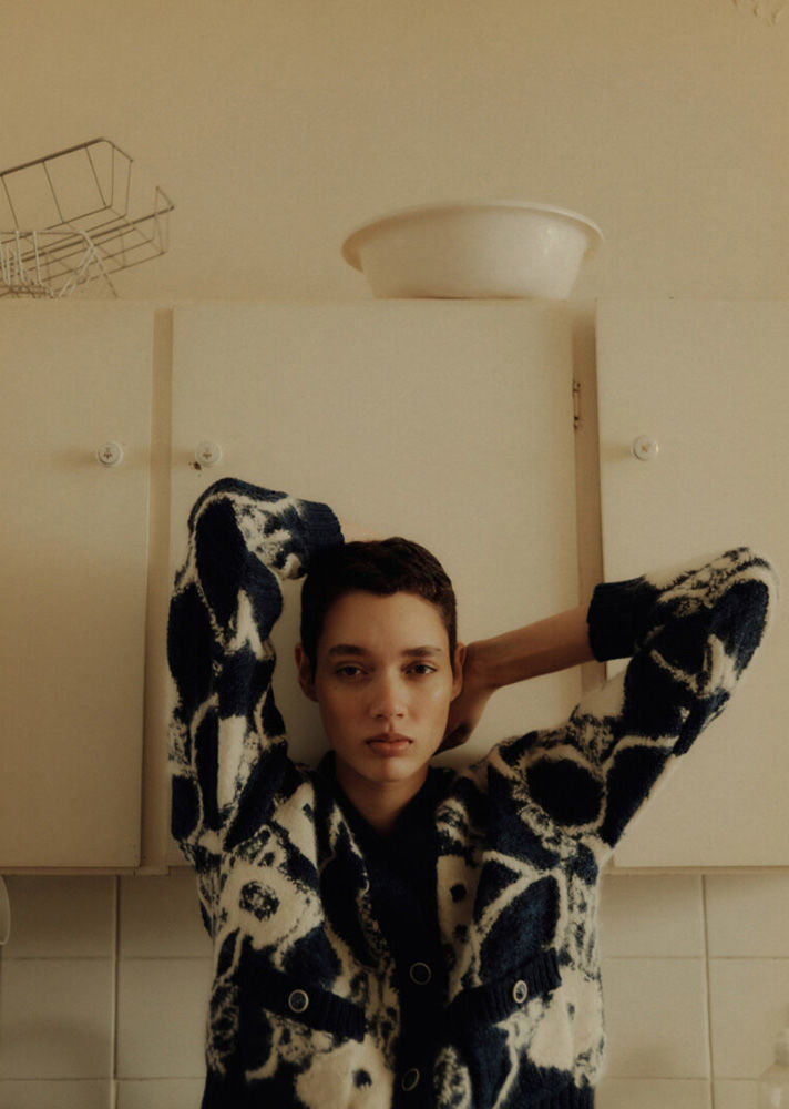 Indoor portrait of a young adult woman with short dark hair, wearing a patterned cardigan, posing against a white kitchen cabinet.