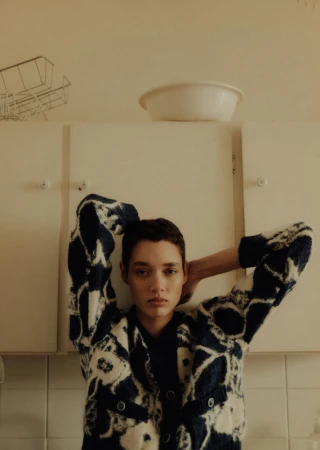 Indoor portrait of a young adult woman with short dark hair, wearing a patterned cardigan, posing against a white kitchen cabinet.