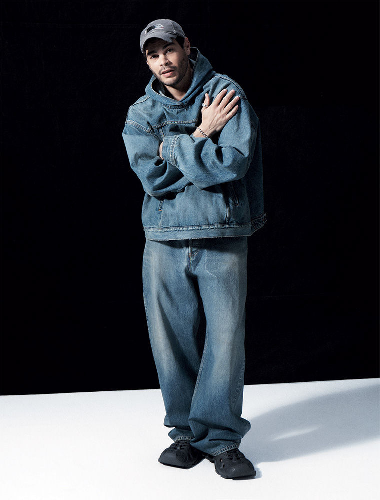 Full-length portrait of an adult male wearing a denim hoodie and pants, standing against a dark backdrop in a photo studio.