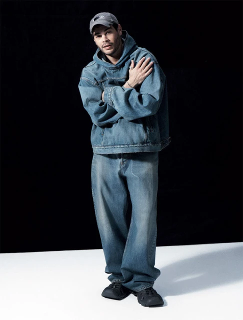 Full-length portrait of an adult male wearing a denim hoodie and pants, standing against a dark backdrop in a photo studio.