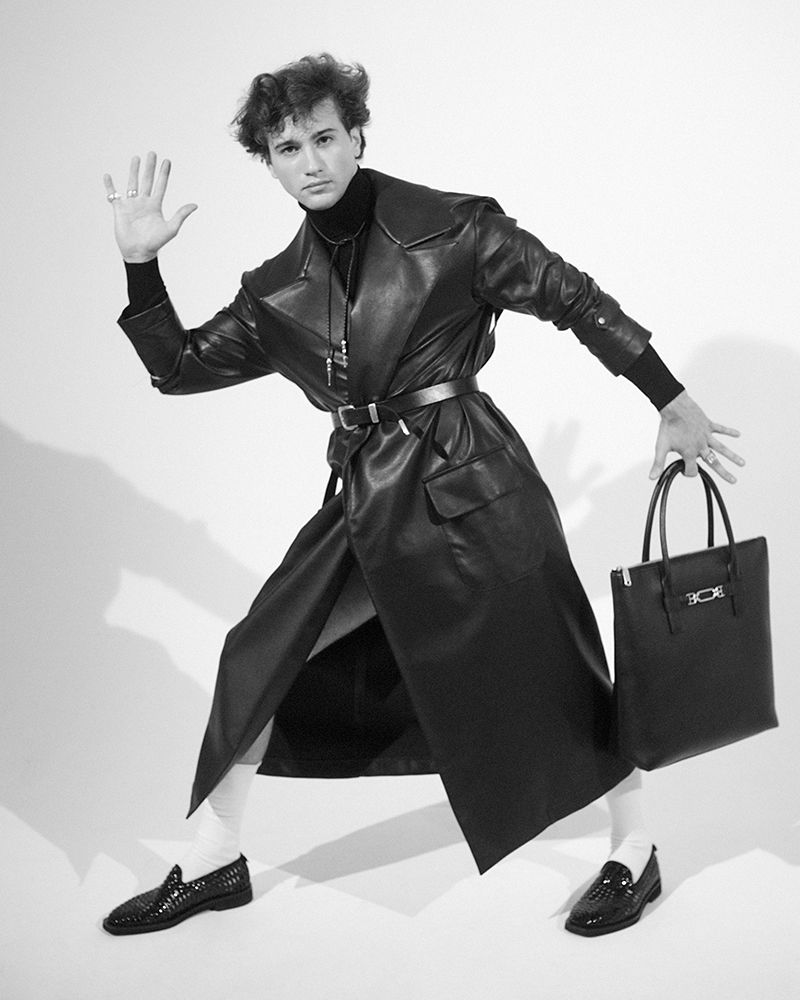 Monochrome fashion shot of a man in a leather trench coat holding a tote bag, striking a pose in a studio setting.