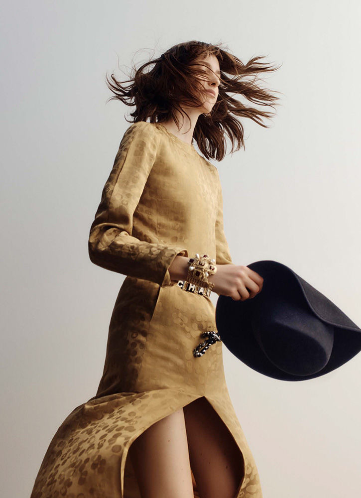 An adult female with wind-blown brunette hair in a patterned tan dress holding a black hat against a light background in a studio shot.