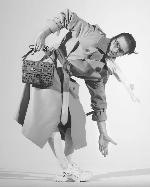 Monochrome fashion shot of a person in a trench coat holding a purse, wearing athletic shoes, standing in a brightly lit white studio.