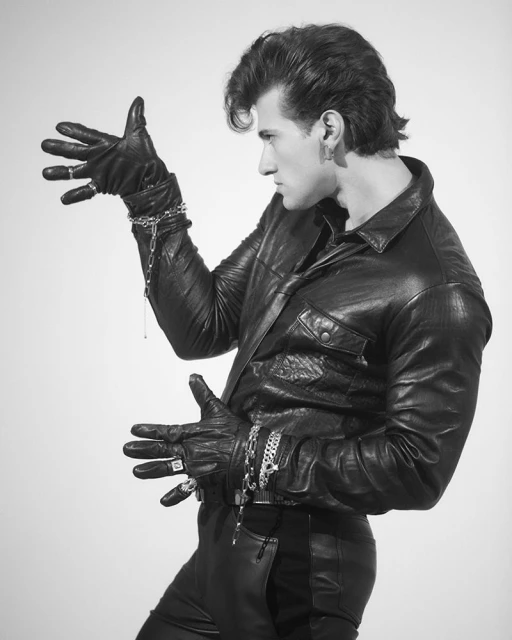 Monochrome portrait of a man in a black leather outfit with metal chains and rings, posing in front of a neutral backdrop. 
