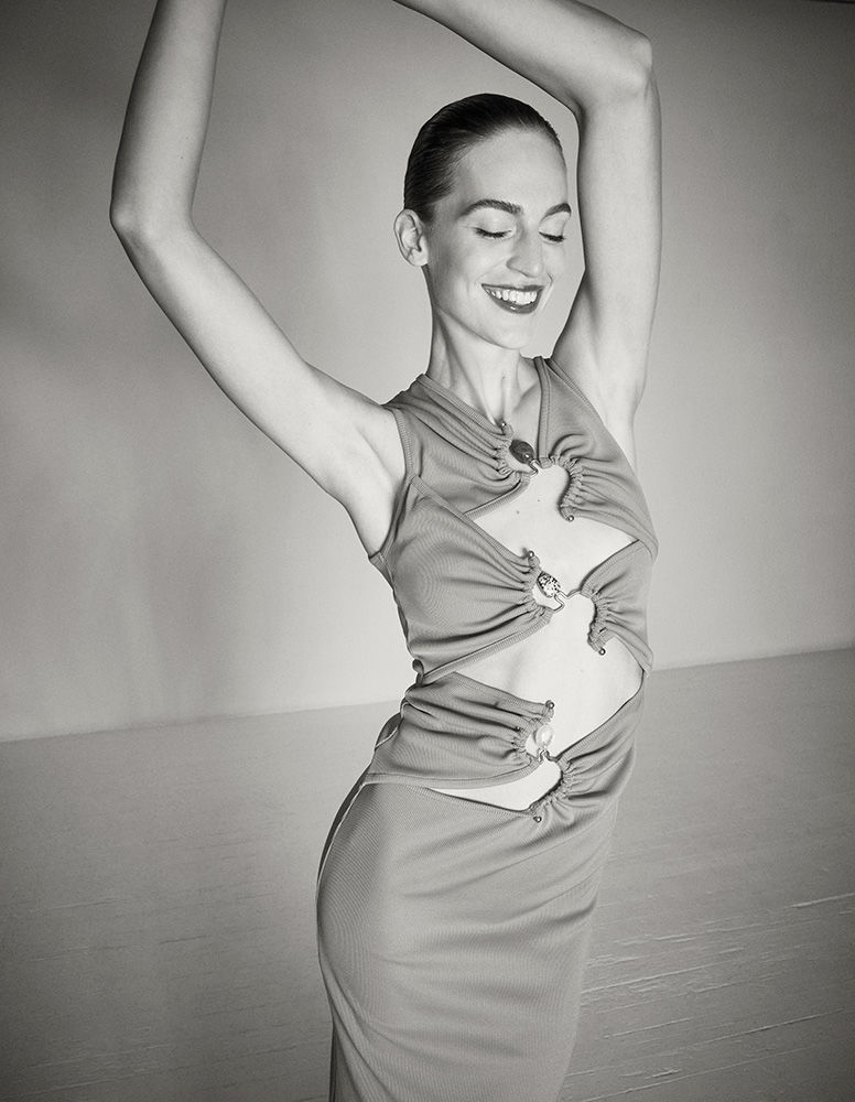 Model with slicked-back hair wearing a cut-out sleeveless dress with raised arms against a solid background