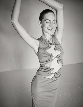 Model with slicked-back hair wearing a cut-out sleeveless dress with raised arms against a solid background