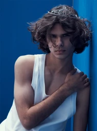Young man with curly brown hair wearing a rhinestone tiara and a white tank top against a blue background
