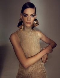 A model with slicked-back dark hair wearing a metallic gold ribbed top and large sculptural gold hoop earrings against a dark backdrop.