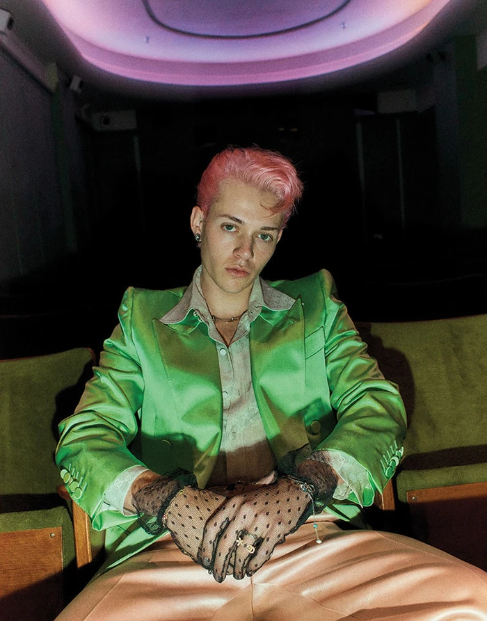 Young man with pink hair wearing a bright green blazer and black lace gloves seated in a dark cinema.