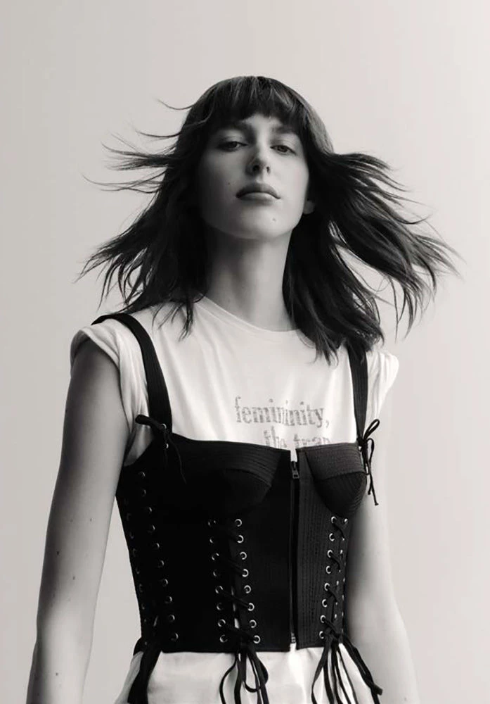 Monochrome portrait of a woman with dark hair and bangs wearing a laced black corset over a white t-shirt against a light background