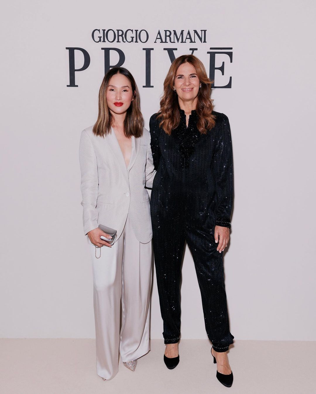 Two women pose in stylish outfits in front of Giorgio Armani Privé backdrop, showcasing fashion and elegance at an event.
