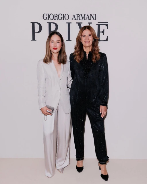 Two women pose in stylish outfits in front of Giorgio Armani Privé backdrop, showcasing fashion and elegance at an event.