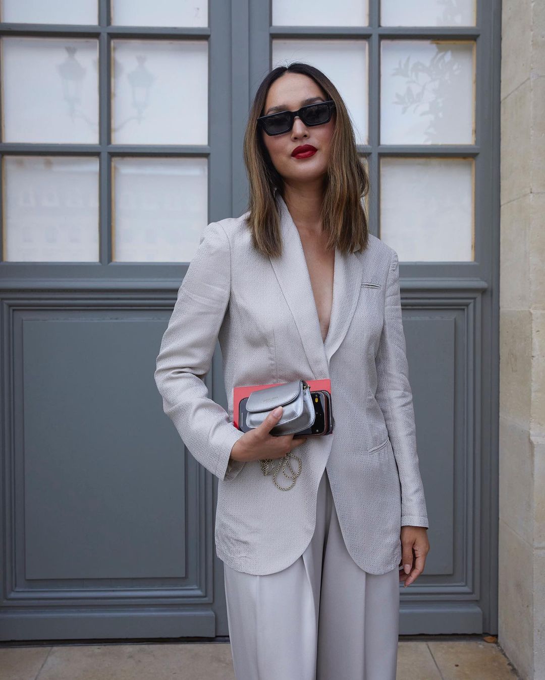 Woman in tailored suit and sunglasses posing against a gray paneled wall, holding clutch; chic and modern fashion photography.