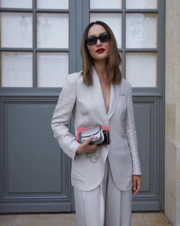 Woman in tailored suit and sunglasses posing against a gray paneled wall, holding clutch; chic and modern fashion photography.