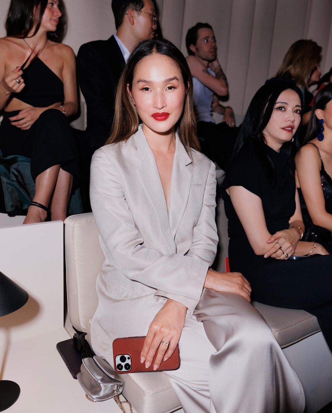 Woman in a light grey suit sits in an indoor space with other people. She is holding a cell phone and wearing bright red lipstick.