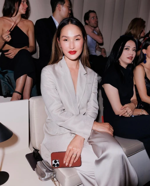 Woman in a light grey suit sits in an indoor space with other people. She is holding a cell phone and wearing bright red lipstick.