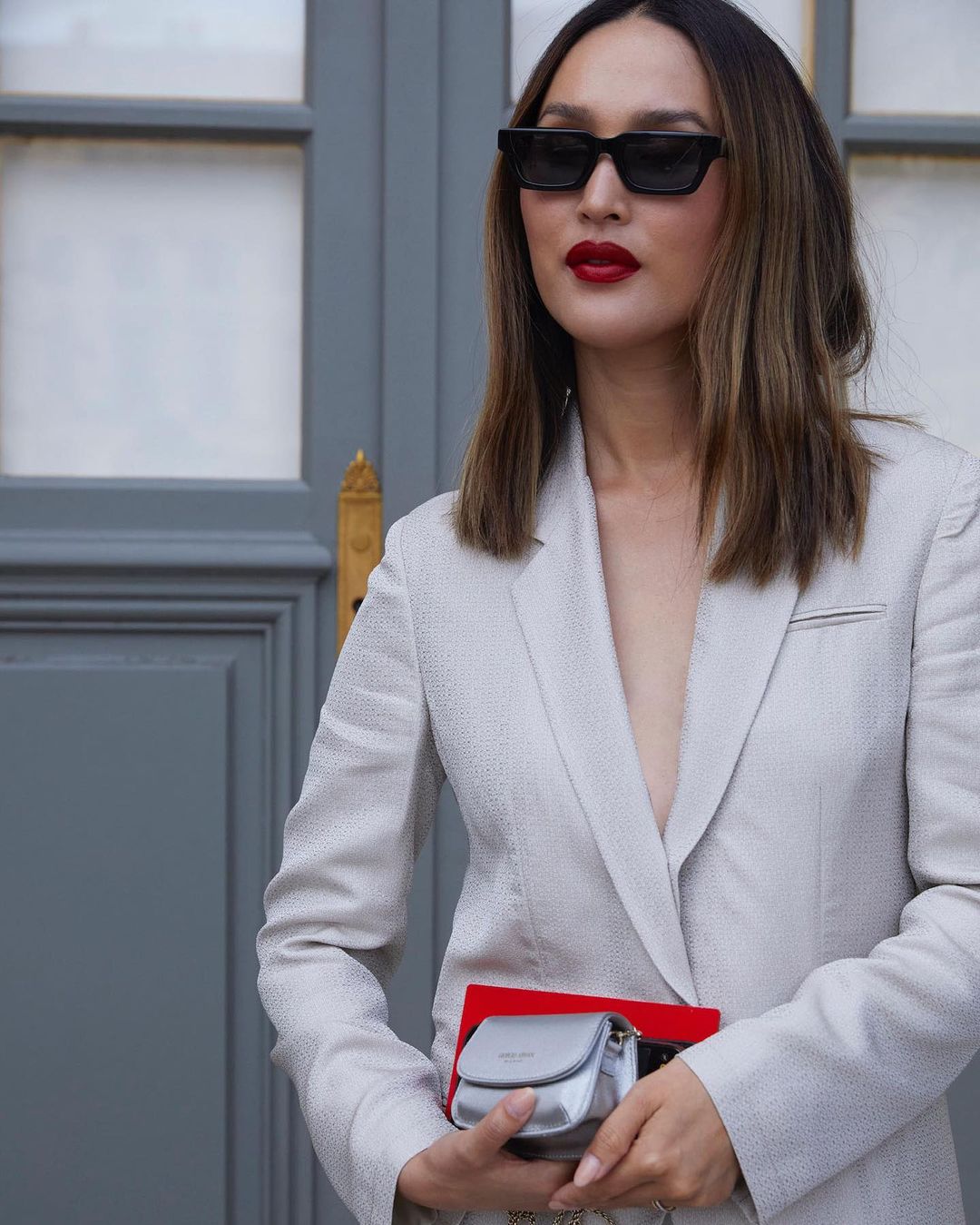 Adult woman in a white suit with sunglasses and red lipstick stands outdoors, holding a small silver purse in a medium shot.