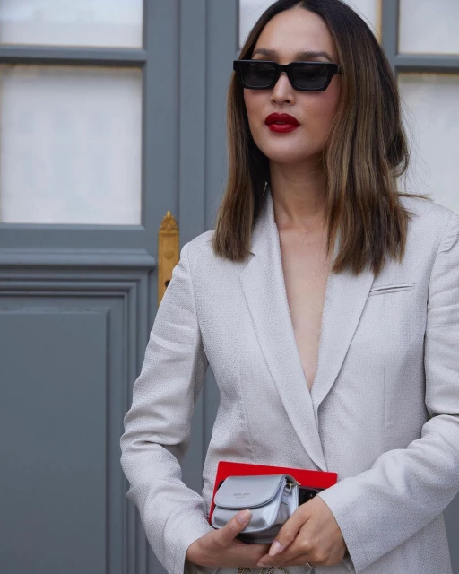 Adult woman in a white suit with sunglasses and red lipstick stands outdoors, holding a small silver purse in a medium shot.