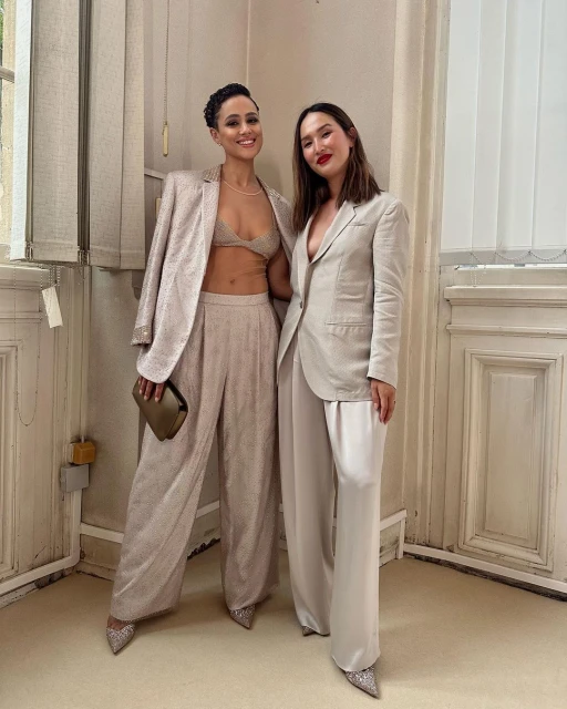 Two adult women in stylish, formal outfits pose indoors, one in a sequined suit, the other in a light-colored blazer and pants.