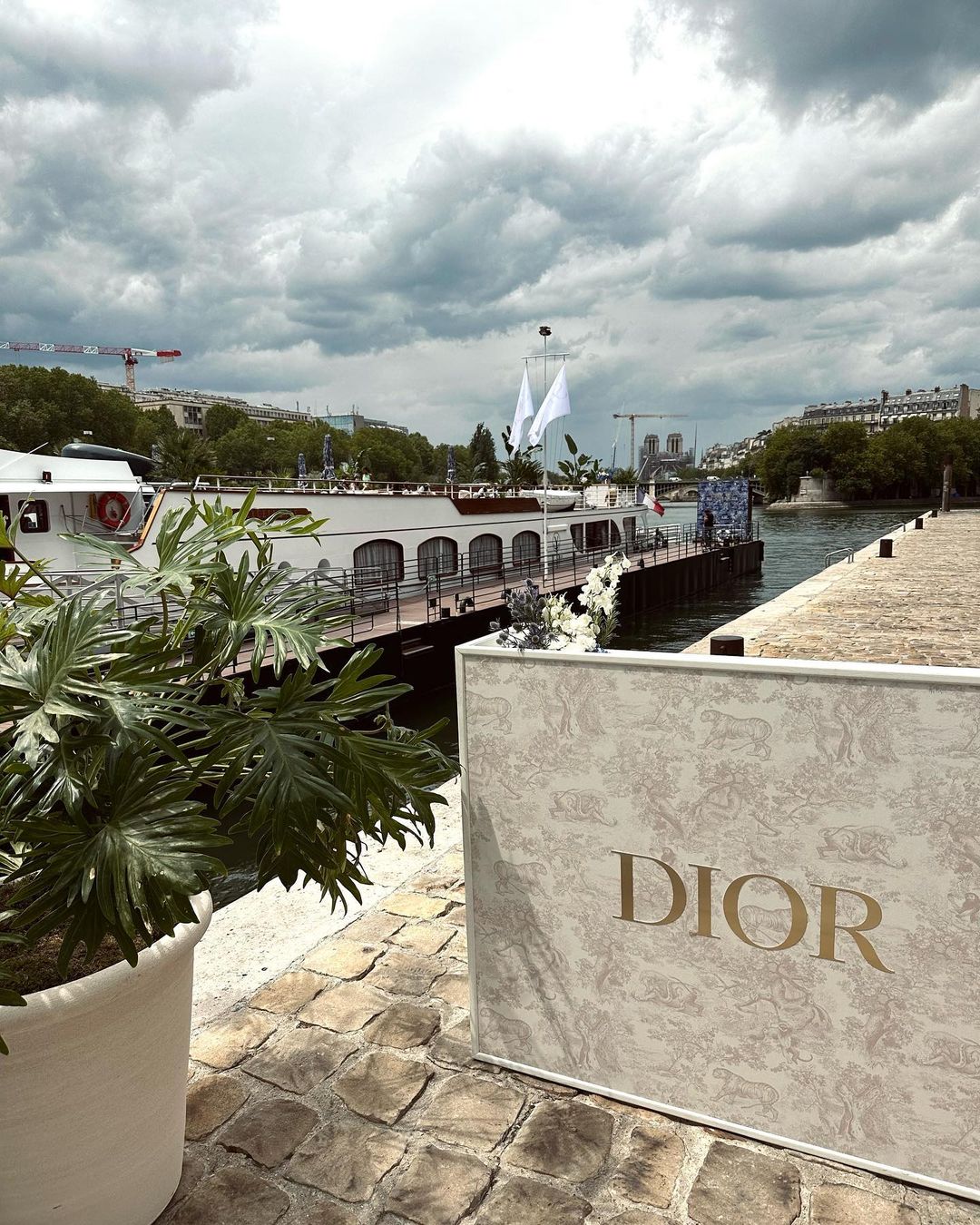 Dior-branded event setup near a river with a docked boat, plant in a pot, and cloudy sky above the cityscape in the background.