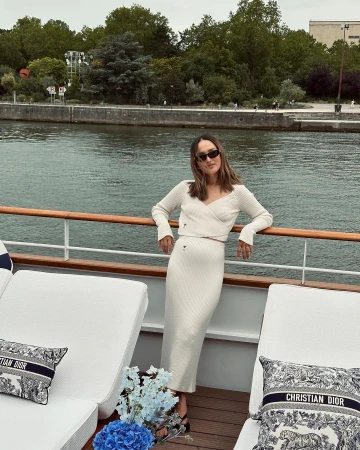 Woman in knitwear poses on a boat with Christian Dior pillows and blue flowers. Water and trees visible in the background.