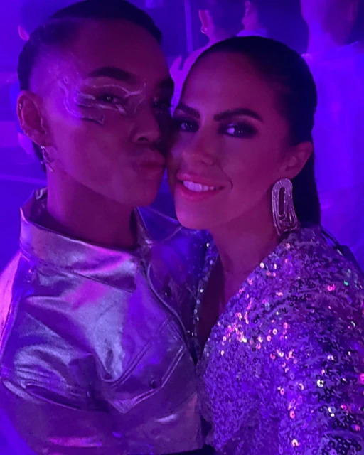 Close-up of a smiling couple at a party: a woman in a sequined dress and a person with makeup wearing a metallic jacket.