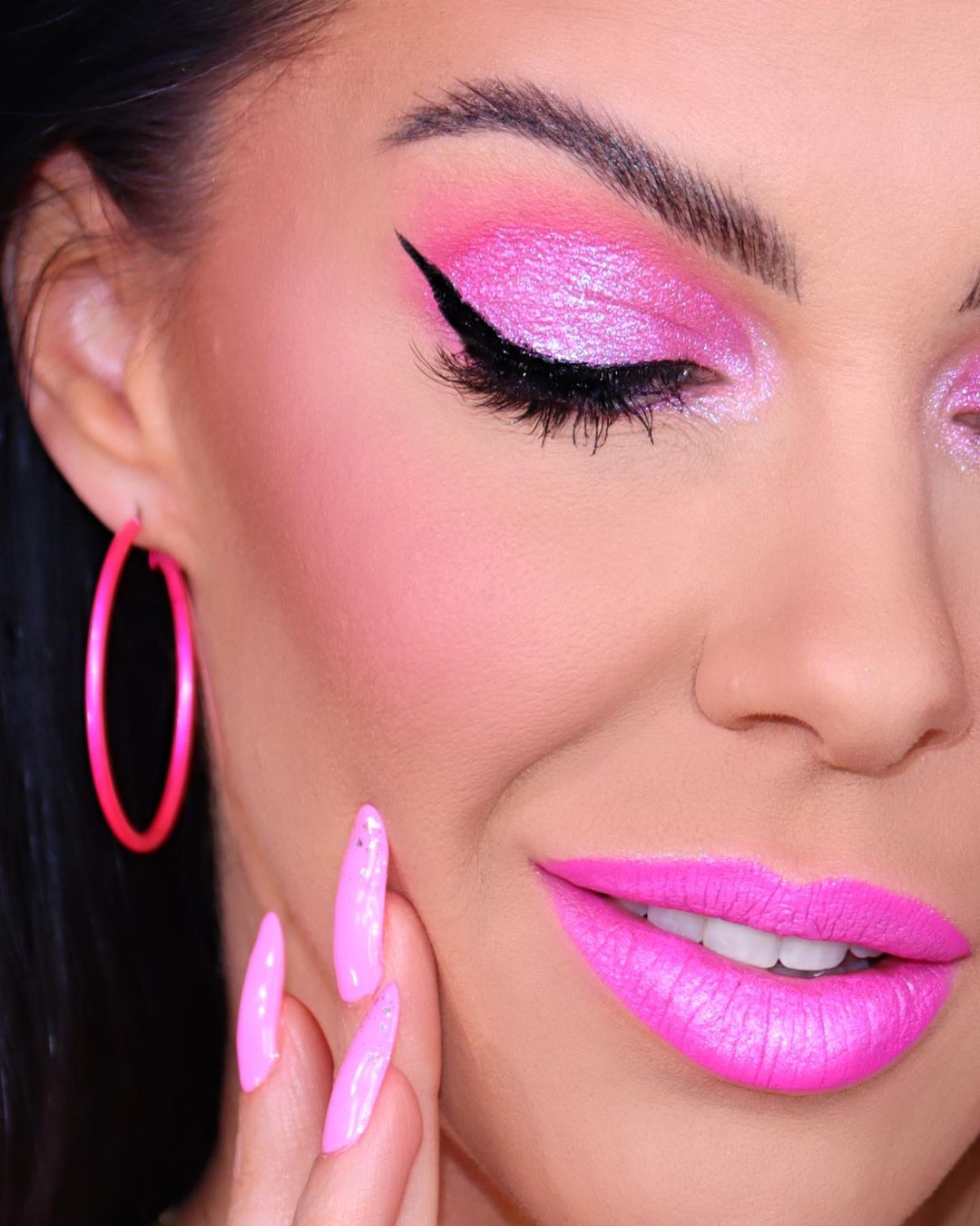 Close-up of a woman with bright pink eye shadow, winged eyeliner, lipstick, and hoop earrings, showcasing makeup and beauty.