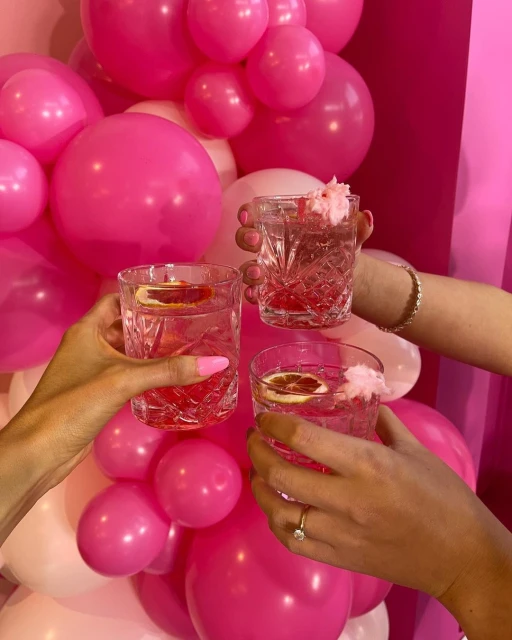 Three cocktails with pink liquid and fruit garnish in crystal glasses held up over a background of pink and white balloons.
