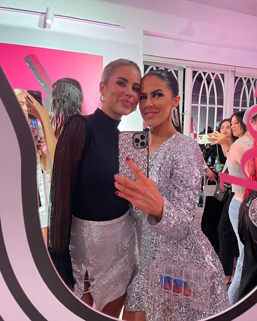 Two adult women posing in front of a mirror in a brightly lit room; one woman wears a silver dress, and the other is in black top and skirt.