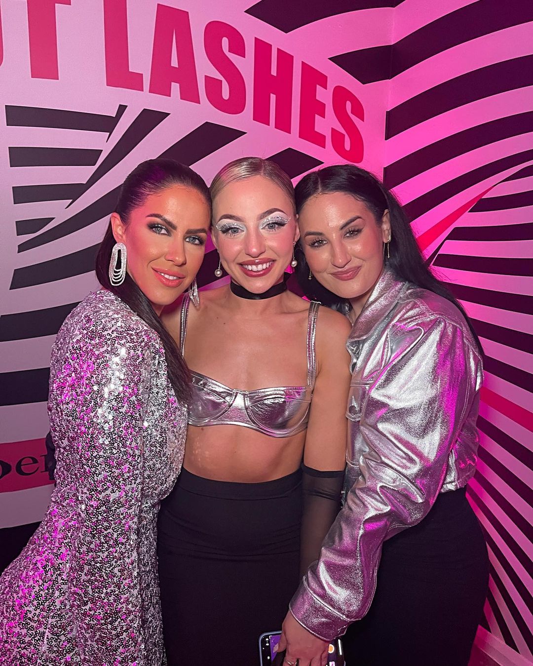 Three adult women pose together in front of a pink and white patterned wall, showcasing sequin and metallic fashion in an indoor setting.