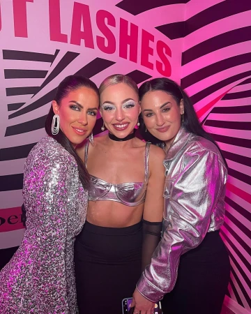Three adult women pose together in front of a pink and white patterned wall, showcasing sequin and metallic fashion in an indoor setting.