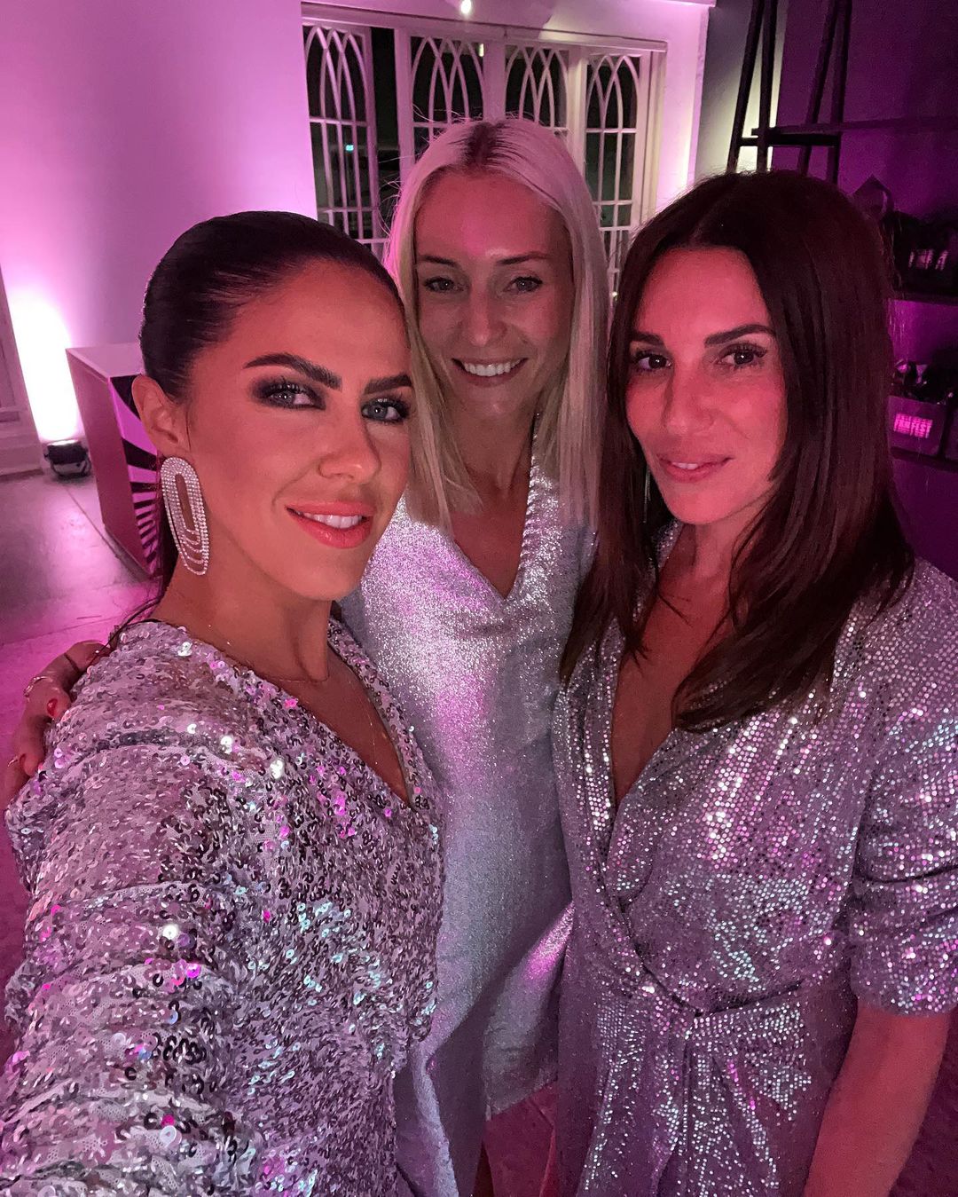 Three women in sparkling dresses pose indoors in a selfie. The photo is lit with a pink hue in a possible party setting.