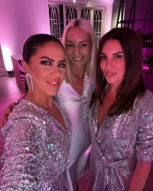 Three women in sparkling dresses pose indoors in a selfie. The photo is lit with a pink hue in a possible party setting.