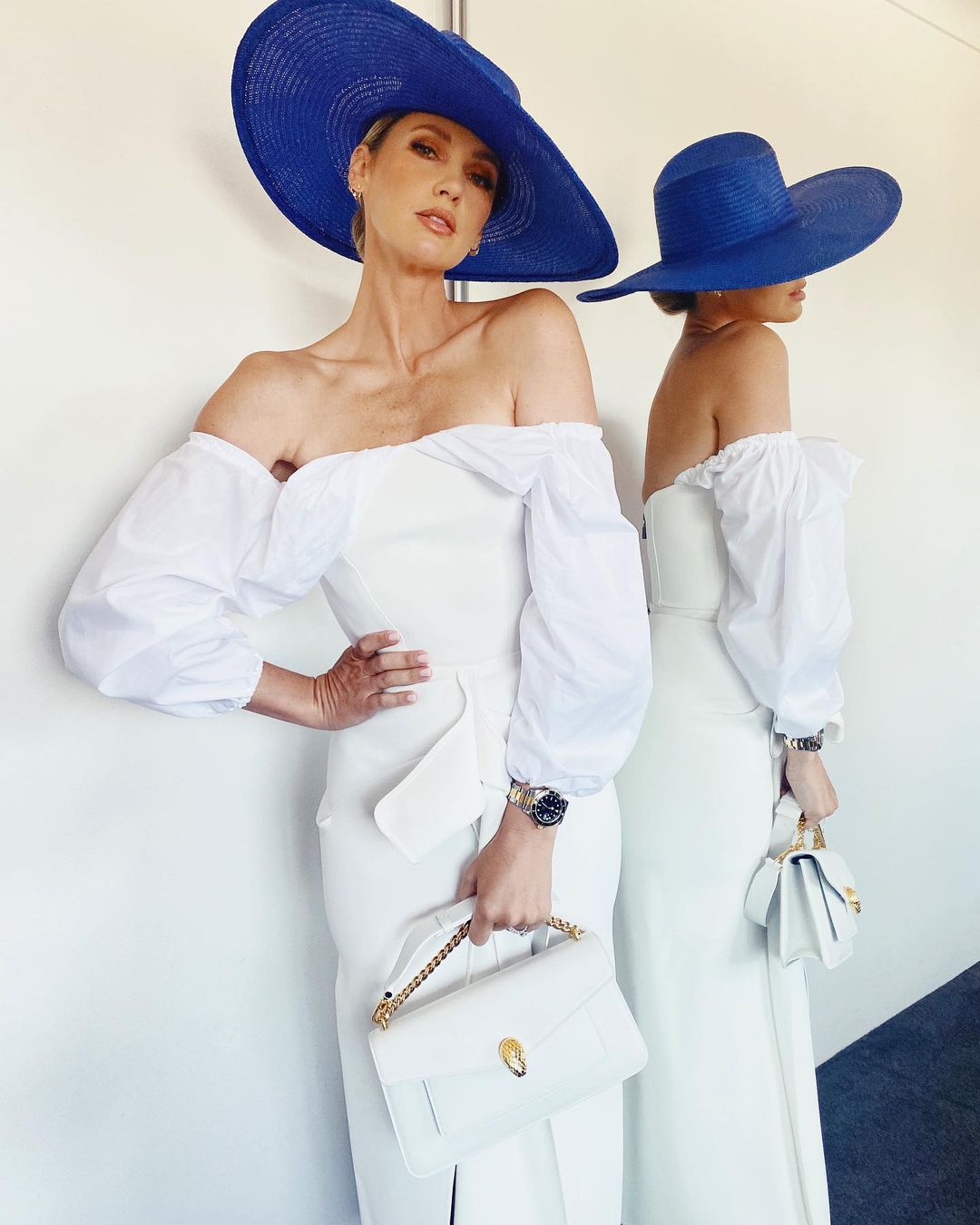 Two women in elegant white dresses and large blue hats pose against a neutral wall. The hats and dresses are the focal point.