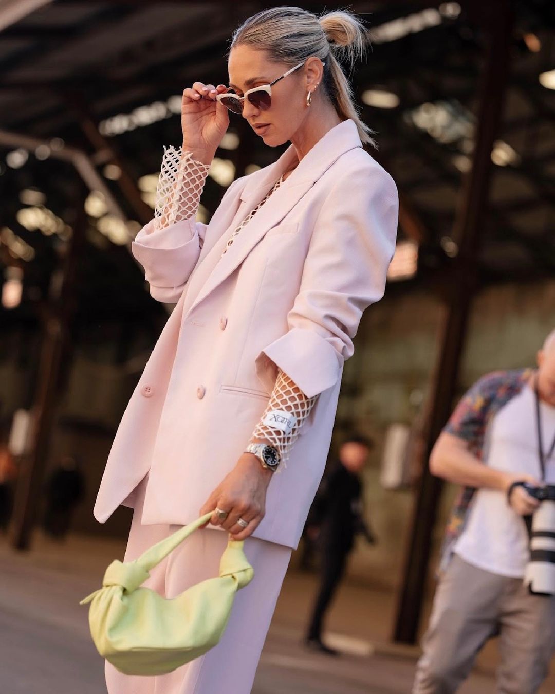 Stylish adult woman poses outdoors in a pink suit, holding a light green handbag and wearing sunglasses; a street style fashion image.