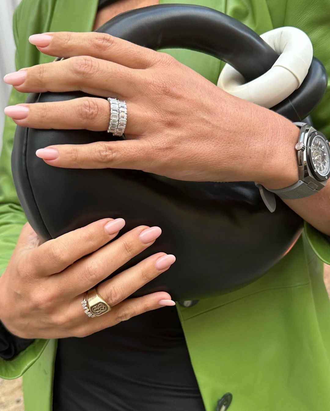 Woman showcasing two diamond rings on her manicured fingers, holding a black purse and wearing a lime green jacket, with a watch.