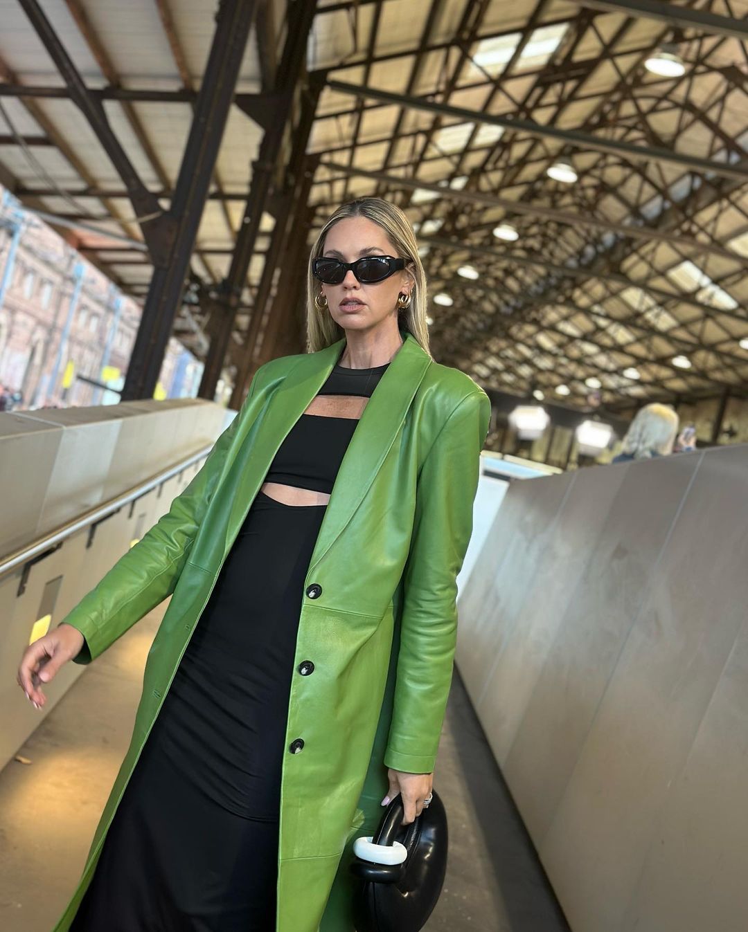 Adult woman with long blonde hair wearing a black dress and a green leather coat, stands in an indoor location with a black handbag.