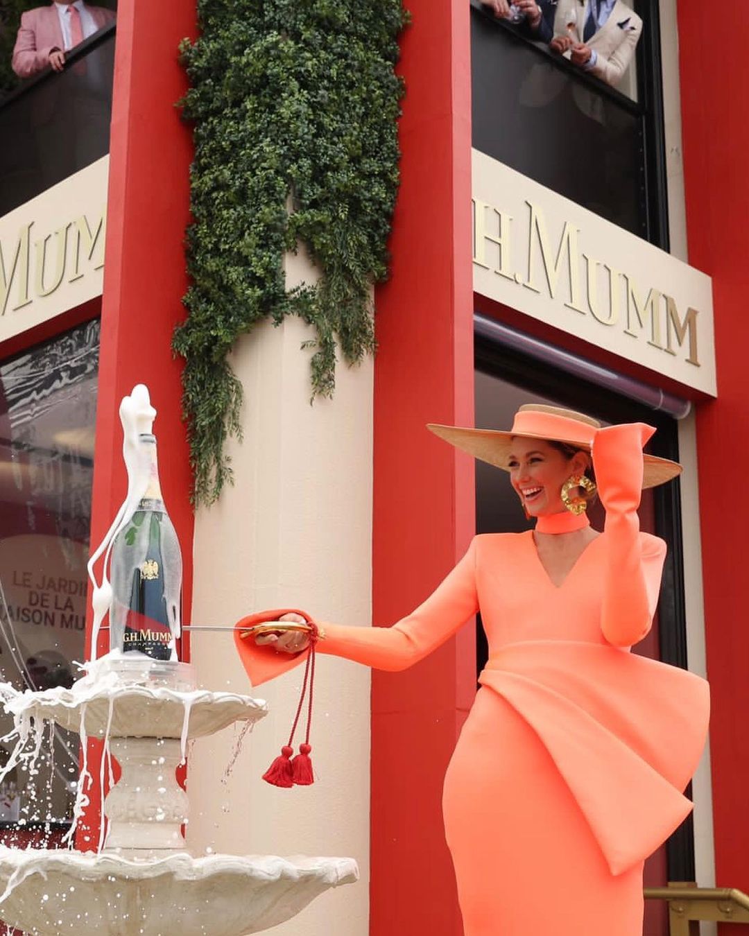 Woman in orange dress opens champagne bottle with sword at an outdoor event, with onlookers in the background on a red building.