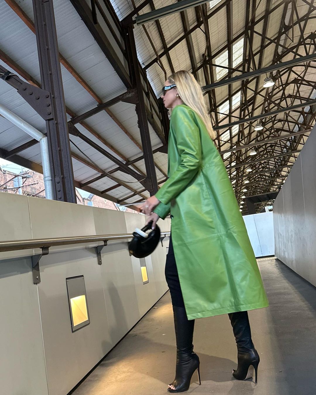 Stylish woman in a vibrant green coat and high-heeled boots walks through an indoor walkway with steel beams and neutral walls.