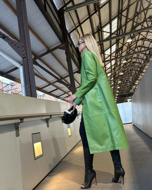Stylish woman in a vibrant green coat and high-heeled boots walks through an indoor walkway with steel beams and neutral walls.