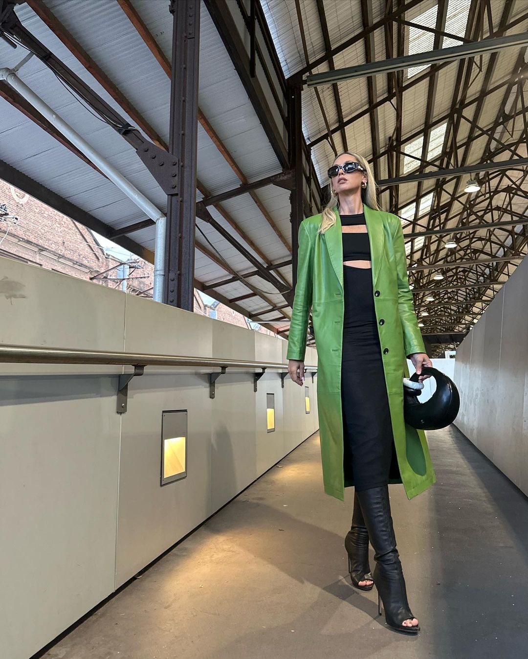 A stylish adult female walks in a light-filled corridor with a green coat, black dress, and handbag, complemented by knee-high stiletto boots.