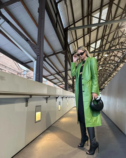 A woman stands under an iron and metal structure wearing a green jacket, a black outfit, sunglasses, and high heeled boots while holding a purse.