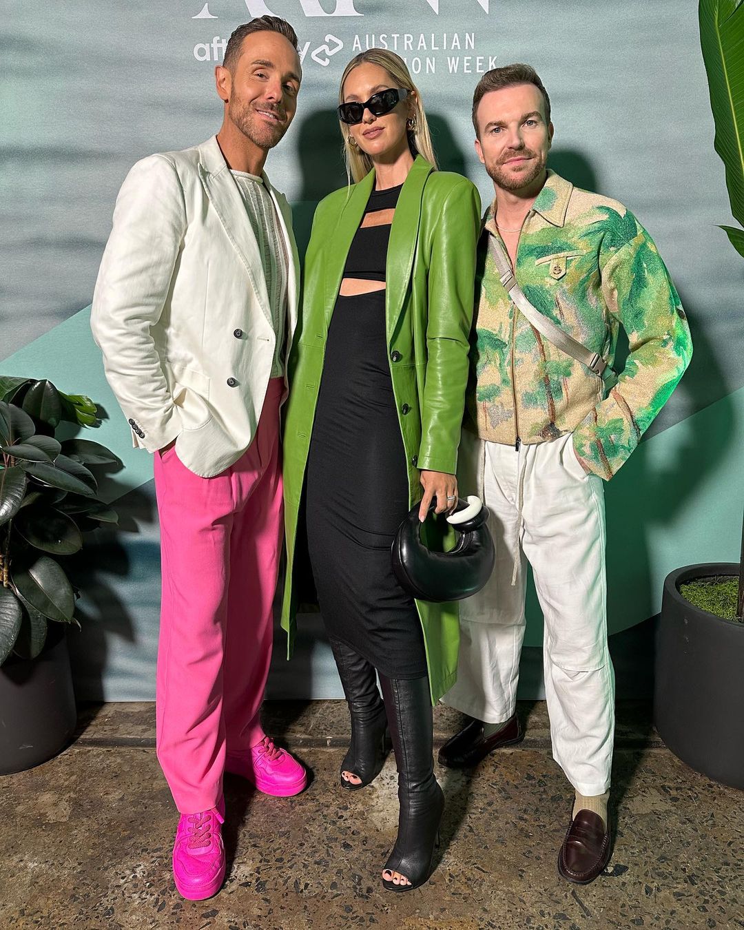 Three people pose together at Australian Fashion Week: two men in white and green shirts and a woman in a green leather coat.